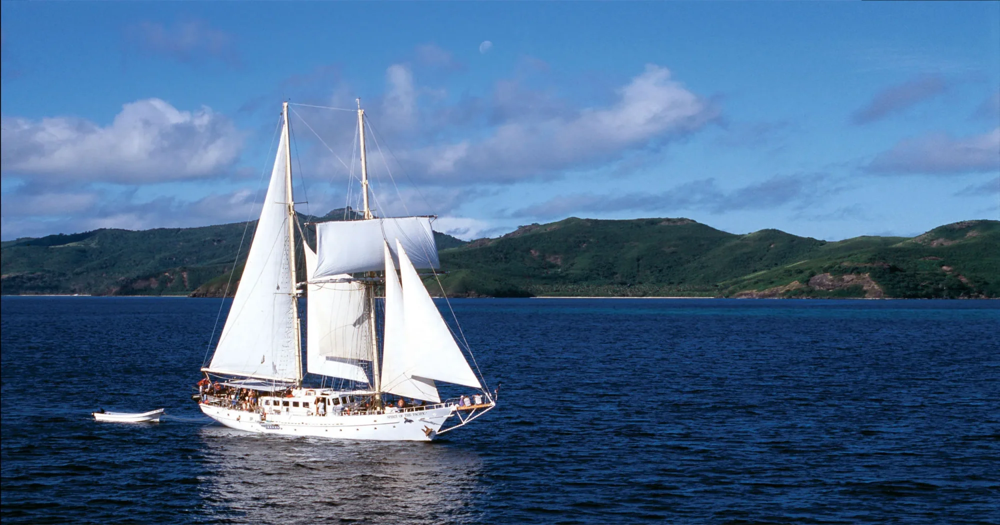 A stunning white-sailed tall ship with traditional rigging anchors in clear blue waters, with lush