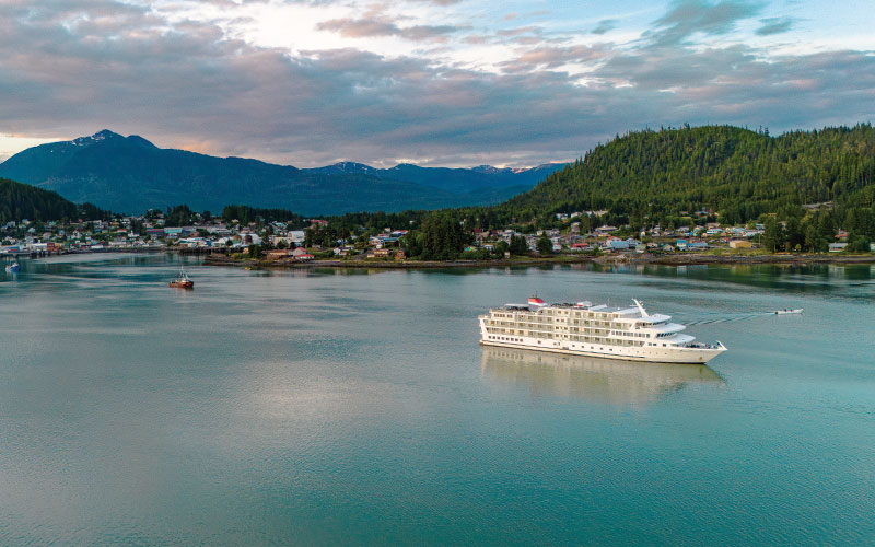 A white cruise ship anchored in a tranquil bay near a forested coastal town with mountains in the background. The calm waters reflect the cloudy sky and surrounding landscape.
