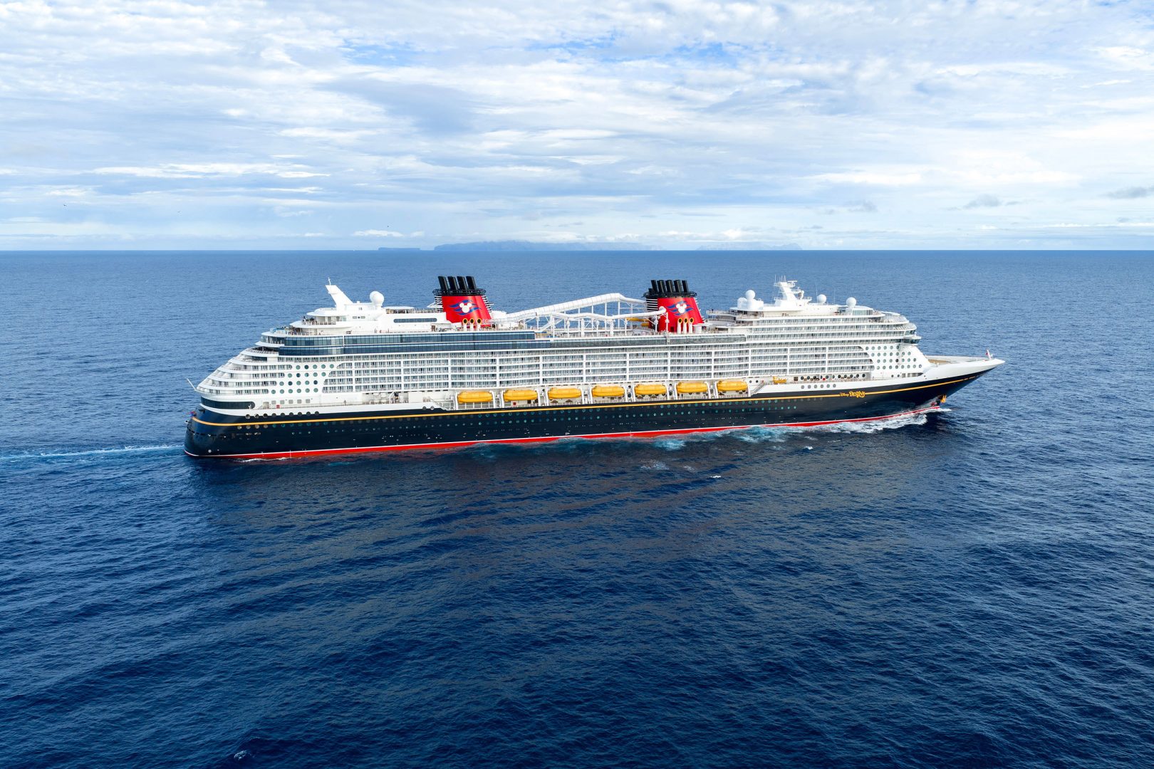 A massive contemporary cruise ship navigates open ocean waters under a partly cloudy sky with distinctive red funnels and dark blue hull.