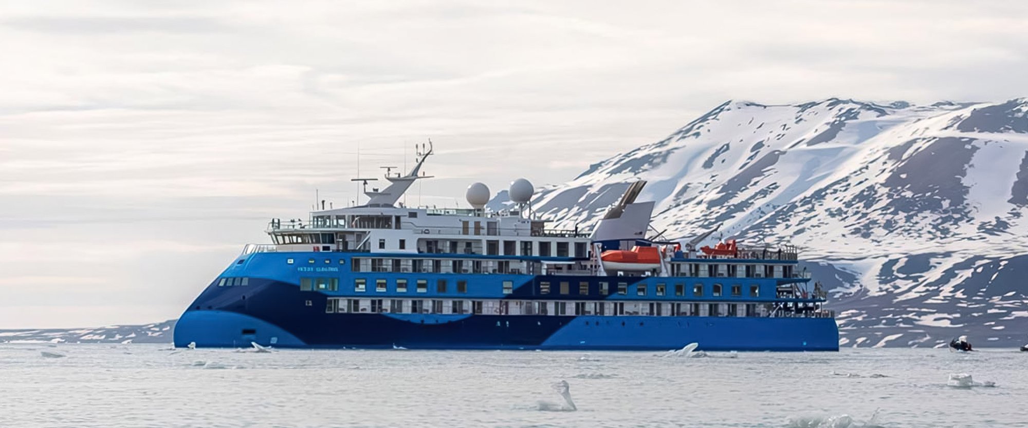 A modern blue expedition cruise ship navigates Arctic waters alongside snow-covered mountains