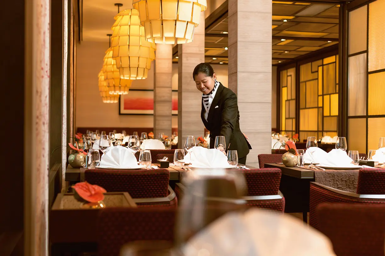 A professional server in formal business attire prepares a fine dining table in an upscale restaurant with warm ambient lighting, white linens, and modern decor.