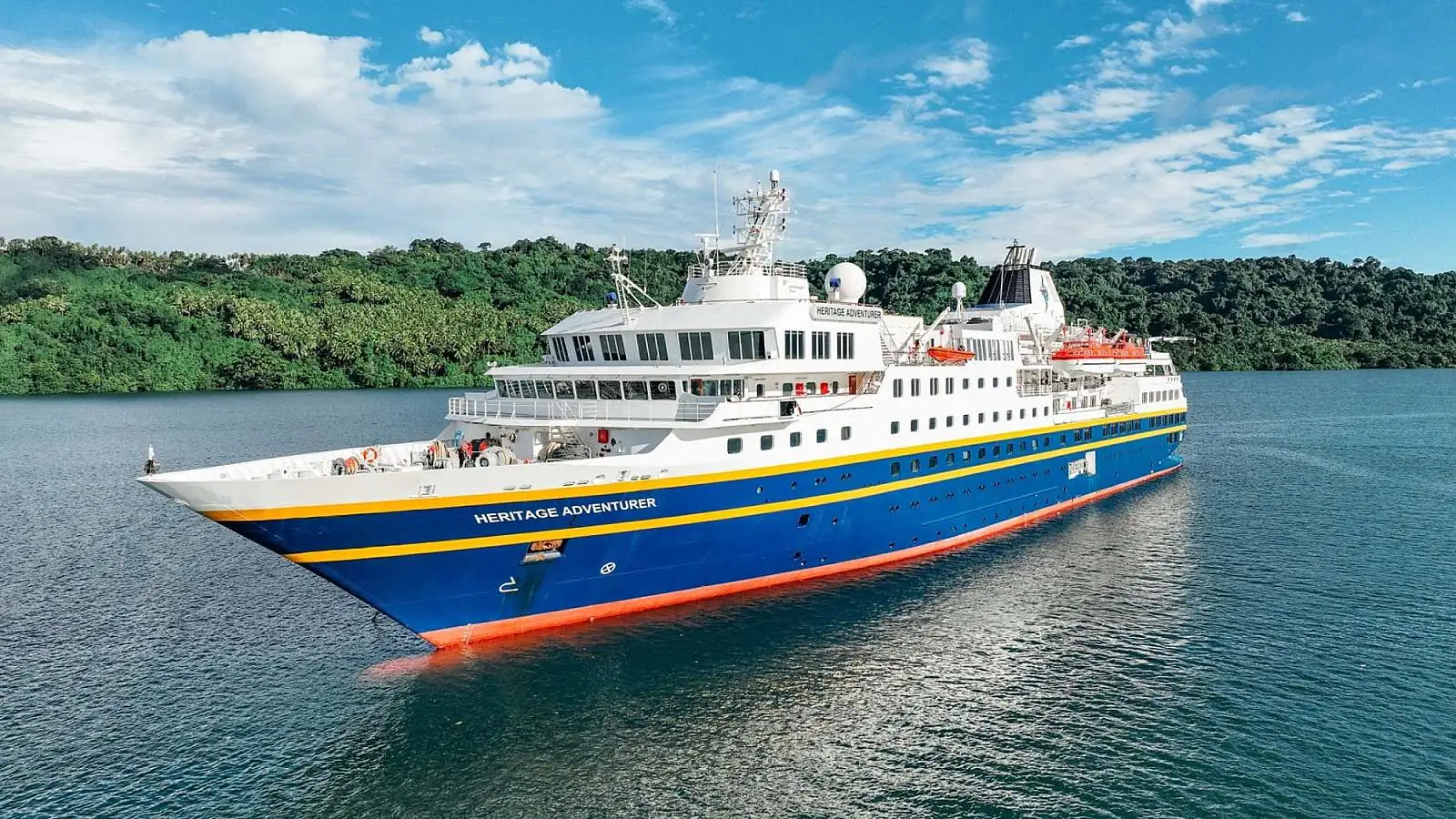 A small expedition-style cruise ship named Heritage Adventurer sailing near a lush green forested