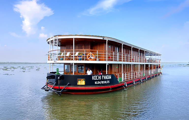 A multi-level wooden river boat named Kochi Pandan floating on calm waters with a blue sky