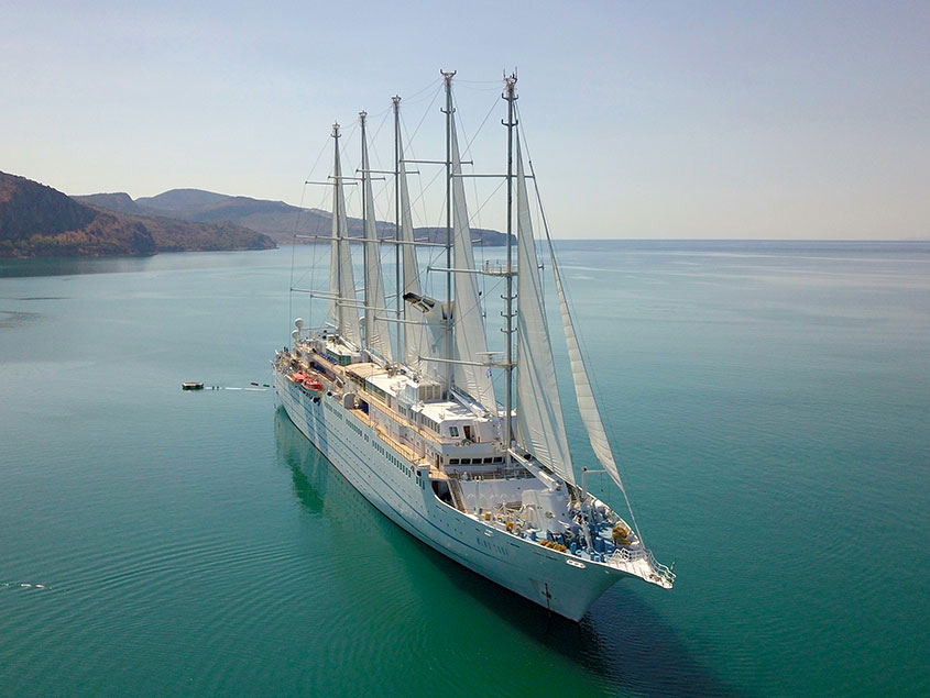 A multi-masted sailing vessel anchored in tranquil blue-green waters with mountainous coastline