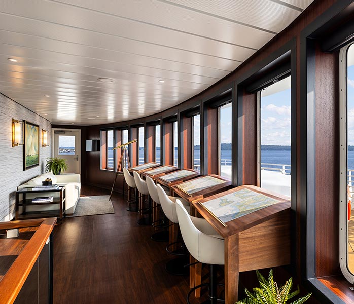 An elegant observation lounge aboard a cruise ship featuring floor-to-ceiling windows overlooking the ocean, with contemporary furnishings including wooden accents, cream-colored chairs, and a sophisticated interior design combining dark wood flooring with white ceiling panels and warm lighting.