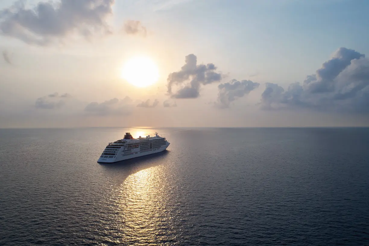 A large modern cruise ship floats peacefully on calm blue ocean waters during golden hour, with