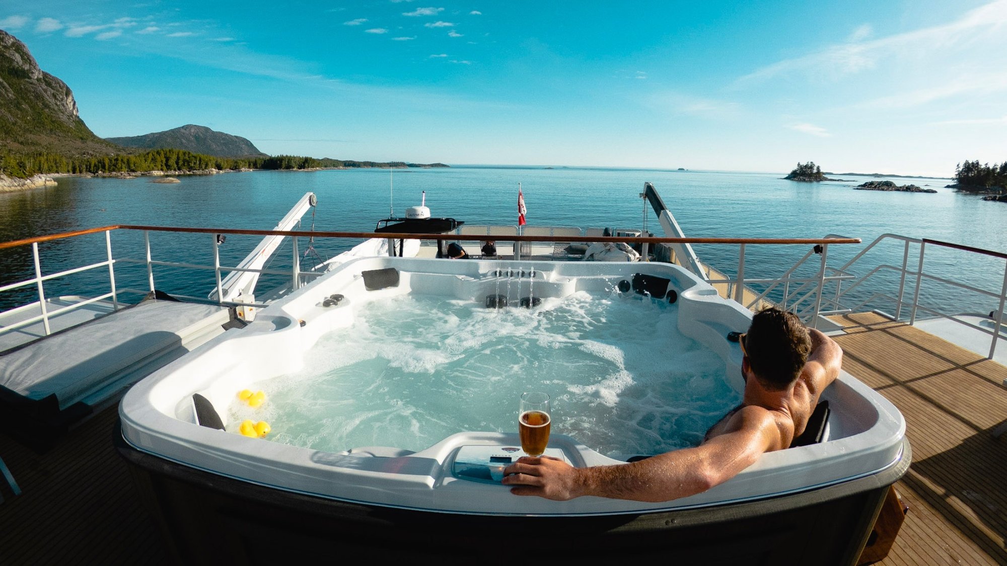 A person relaxes in a large, bubbling hot tub on the deck of a luxury yacht, holding a beverage while enjoying panoramic views of a pristine turquoise lake surrounded by forested mountains and rocky shores.