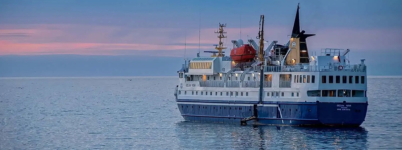 A blue and white expedition cruise ship anchored in calm waters during twilight hours, featuring
