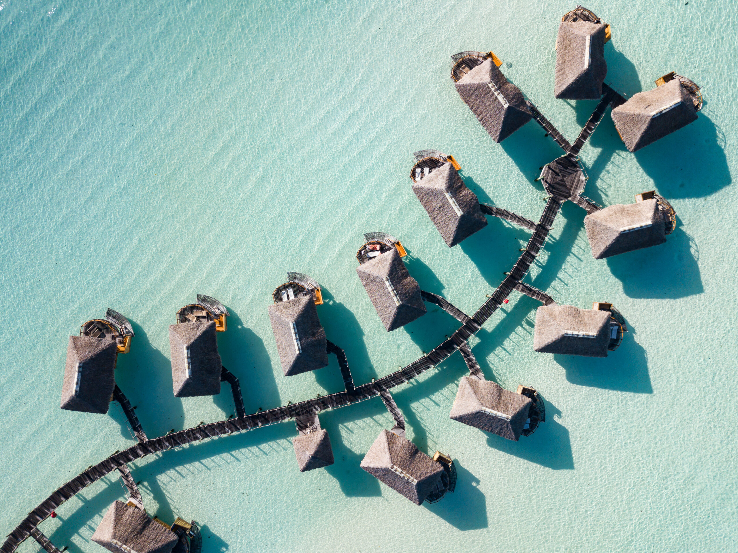An aerial view of a luxury resort with thatched-roof overwater bungalows arranged in a curved pattern along wooden walkways in crystal-clear turquoise lagoon waters.