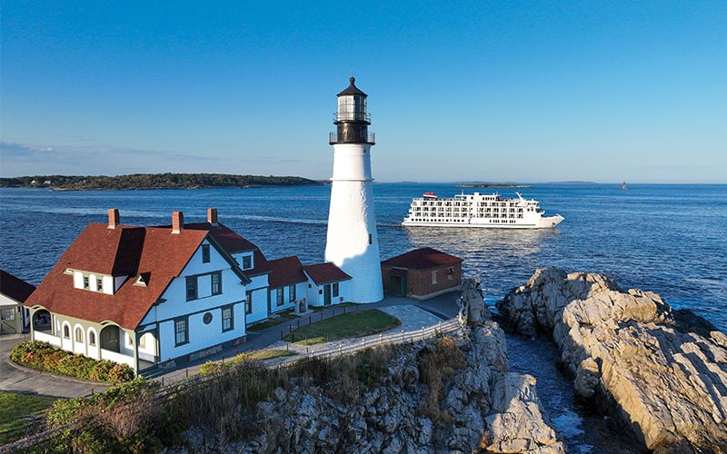 A white lighthouse with a red-roofed keeper's house sits on rocky coastal terrain, with a white