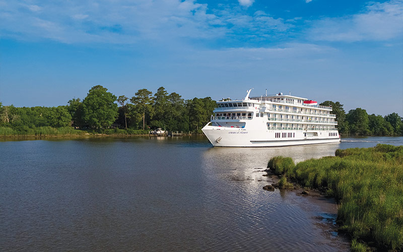 A white river cruise vessel sailing through a serene waterway with lush green trees and grassy shorelines. The ship appears to be a modern, multi-deck river cruise ship navigating a calm, narrow river.