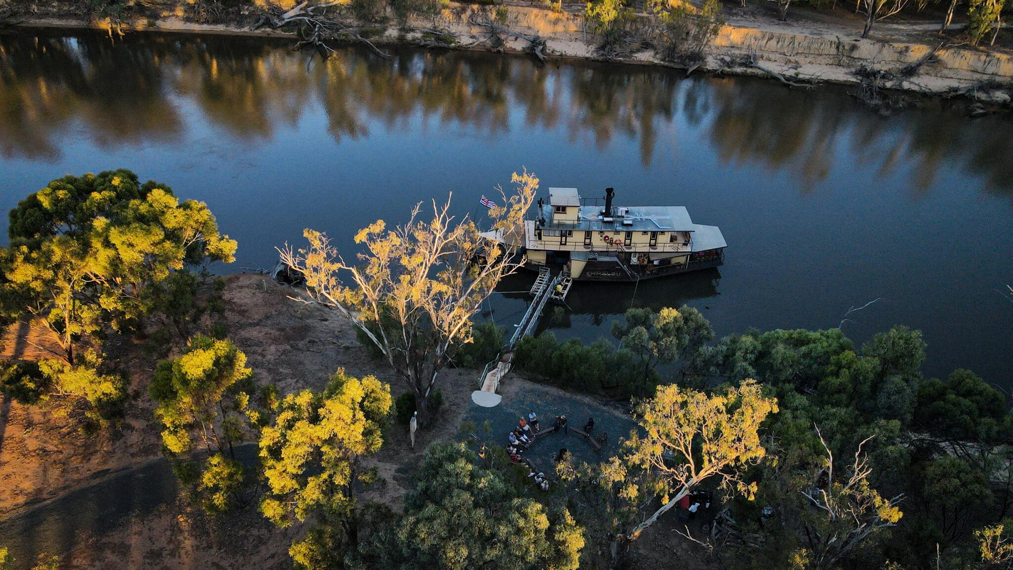 An aerial view of a paddlewheel riverboat moored along the Murray River in Australia, with passengers gathered on the sandy bank near the gangway. Golden-lit eucalyptus trees frame the scene, reflecting in the calm blue water, showcasing a scenic inland waterway cruise experience.