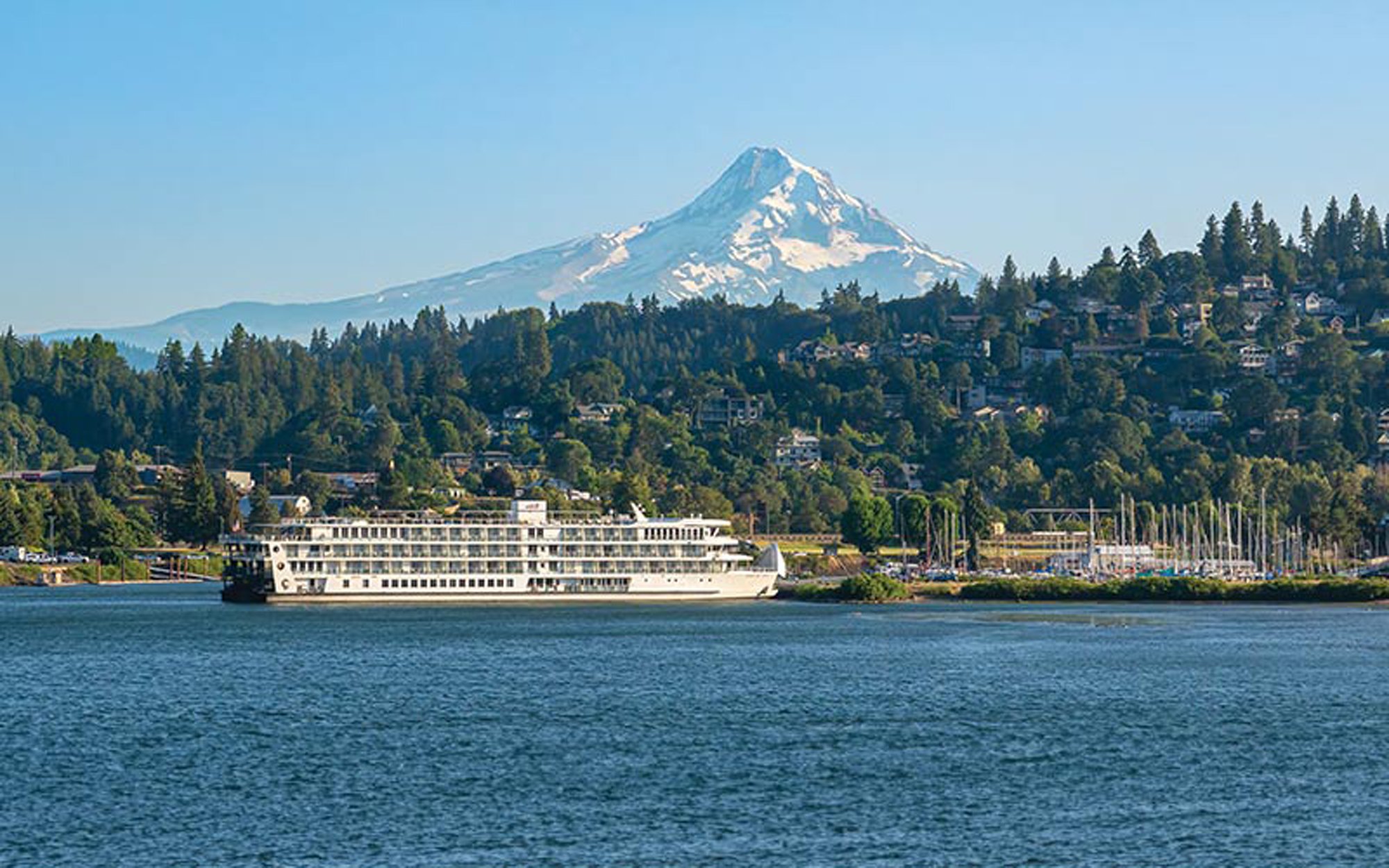 A multi-deck white riverboat navigates calm blue waters with snow-capped Mount Hood dominating the background, surrounded by dense evergreen forests and a hillside community nestled among the trees in the Pacific Northwest.
