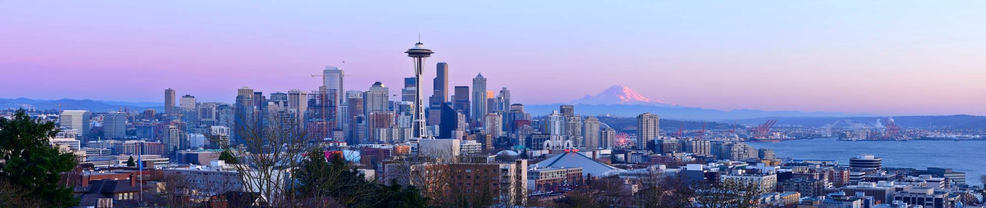 A stunning panoramic view of Seattle's iconic skyline at sunset, featuring the Space Needle as the central landmark, with Mount Rainier visible in the distance and Puget Sound stretching across the foreground.