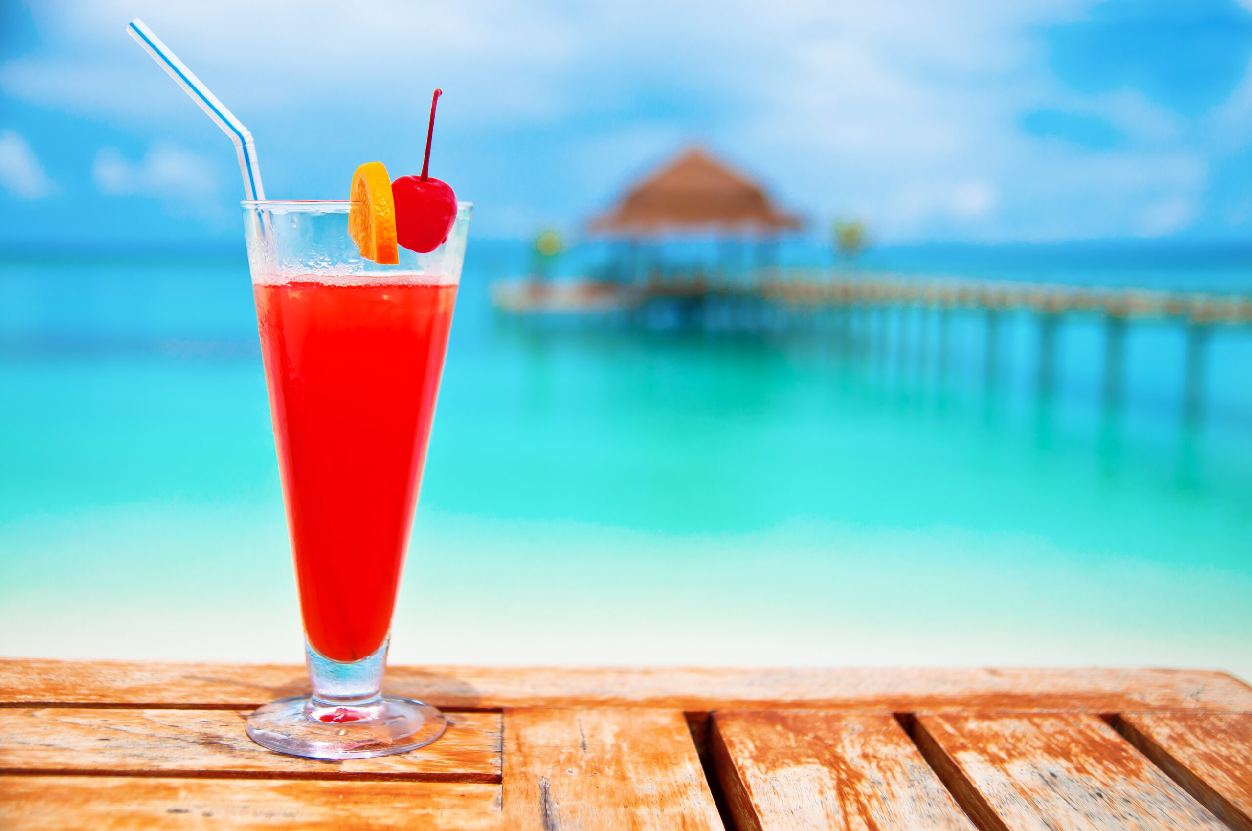 A vibrant red cocktail with ice, garnished with a maraschino cherry and orange slice, sits on a wooden deck overlooking turquoise tropical waters.