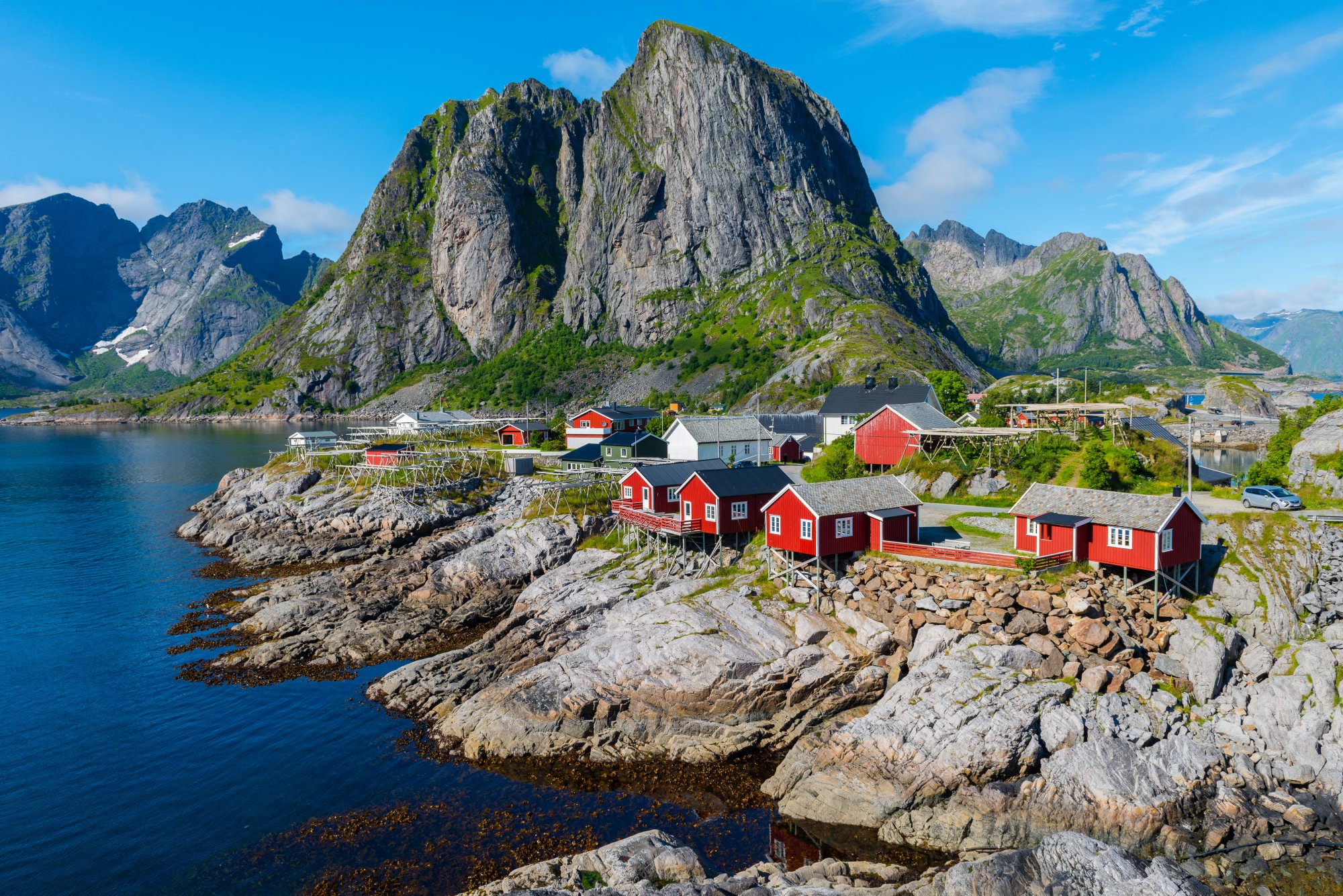 Scenic Norwegian fjord with steep green mountainsides, a small village nestled at the water's edge, and a cruise ship sailing through the calm deep blue waters.