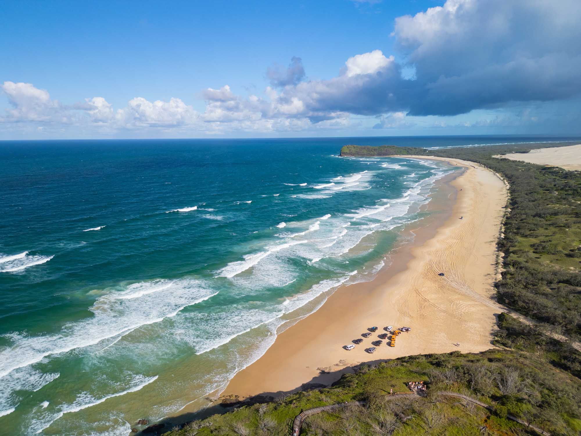 A stunning aerial photograph of 75 Mile Beach on K'gari Island (Fraser Island) in Queensland, Australia, showing a pristine golden sand beach stretching along turquoise ocean waters with white-capped waves, parked vehicles on the beach, lush green vegetation inland, and a dramatic sky with cumulus clouds.