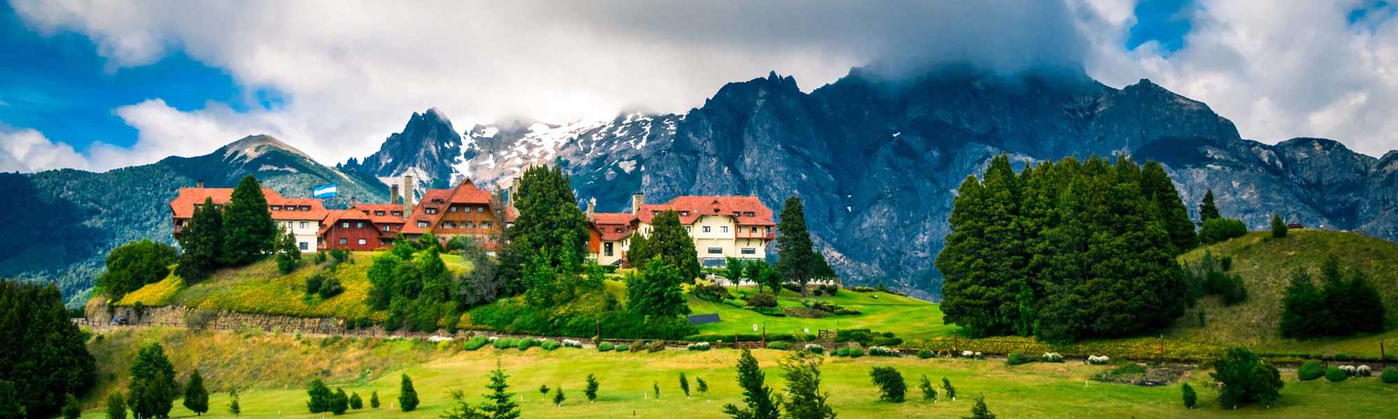 A striking castle with distinctive orange-red roofs nestled on a hillside amid lush green meadows, surrounded by dramatic snow-capped Alpine peaks and dense evergreen forests. The picturesque setting exemplifies classic Swiss alpine architecture and landscape.