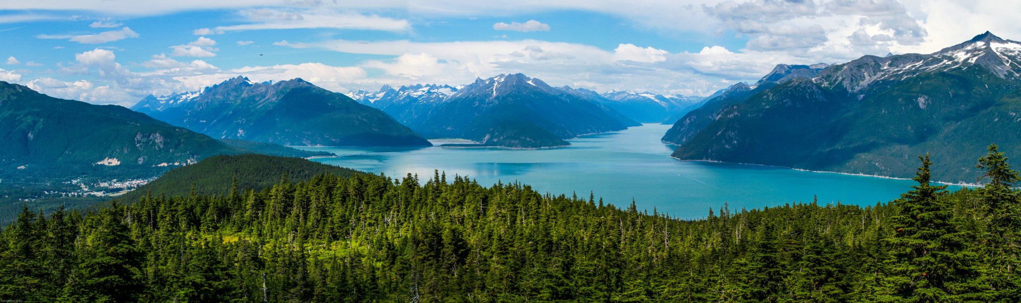 A breathtaking panoramic view of a pristine turquoise alpine lake surrounded by dense coniferous forests and dramatic snow-capped mountain peaks.