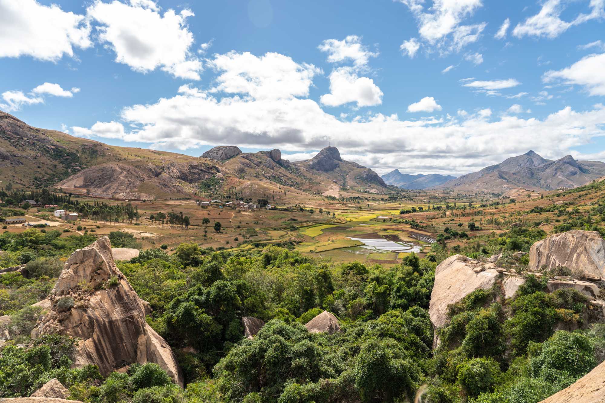 A panoramic view of the Anja Community Reserve in Madagascar, featuring distinctive large granite boulders scattered across a lush green landscape with rice paddies, distant mountains, and a winding river. The scene combines dramatic rocky outcrops with cultivated agricultural fields and natural vegetation under a partly cloudy sky.