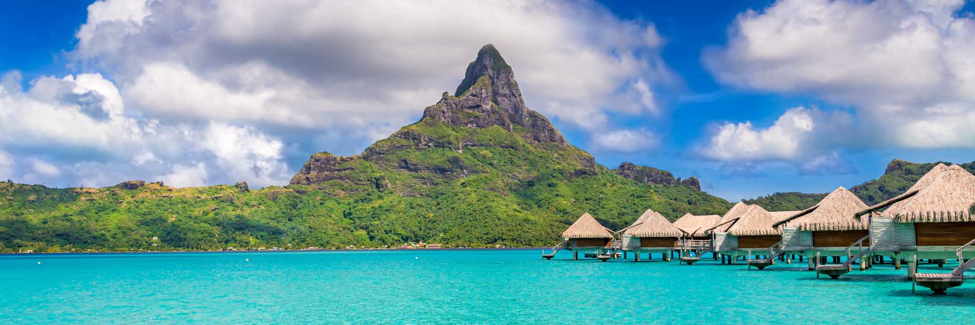 A stunning tropical paradise view of Bora Bora Island in French Polynesia, featuring iconic thatched-roof overwater bungalows in turquoise lagoon waters, with the dramatic volcanic Mount Otemanu peak rising majestically in the background.