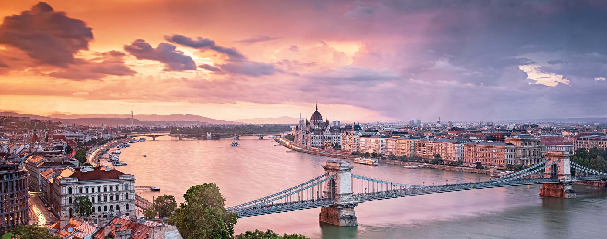 A stunning panoramic view of Budapest at golden hour, featuring the iconic Chain Bridge spanning the Danube River with the Hungarian Parliament Building illuminated on the right bank.