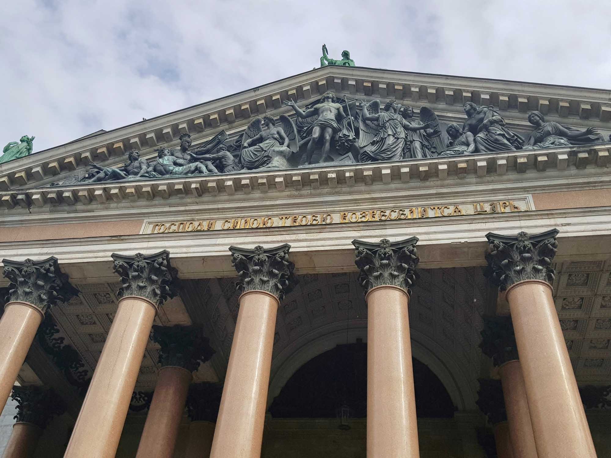 A striking neoclassical facade featuring six tall Corinthian columns with ornate capitals supporting an elaborate pediment adorned with classical sculptural reliefs. The building displays characteristic Beaux-Arts architecture with detailed stonework and Cyrillic inscription text visible above the colonnade.