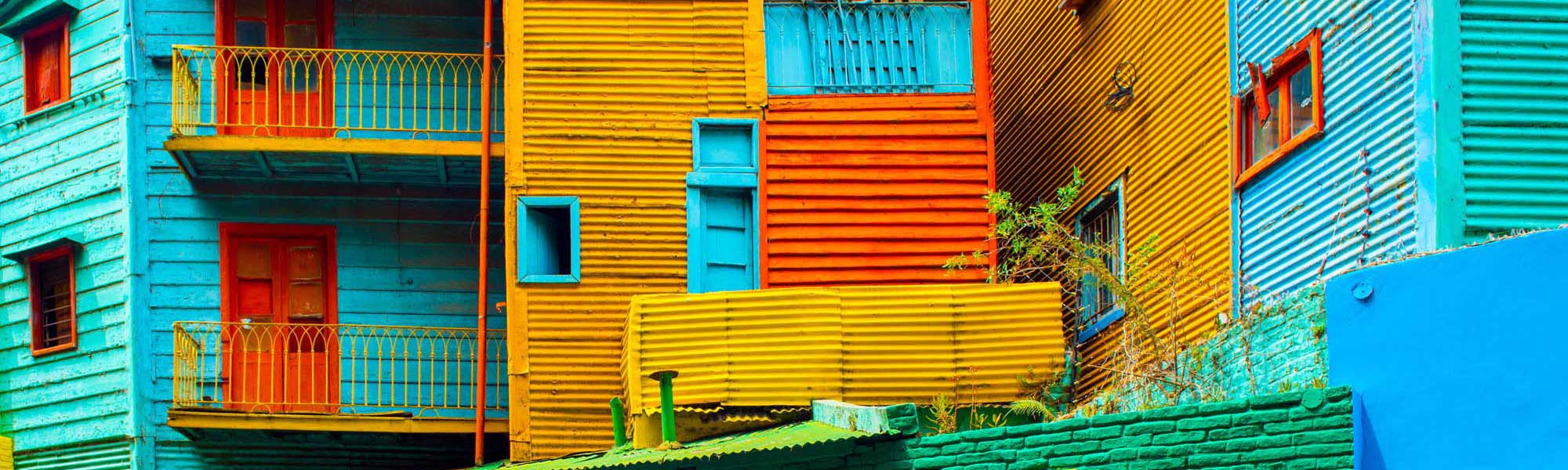 A vibrant street scene featuring iconic brightly painted corrugated metal buildings in shades of turquoise, yellow, orange, and red with decorative iron railings and shuttered windows. This picturesque neighborhood showcases the distinctive architectural style and rich cultural heritage of Buenos Aires' historic La Boca district.