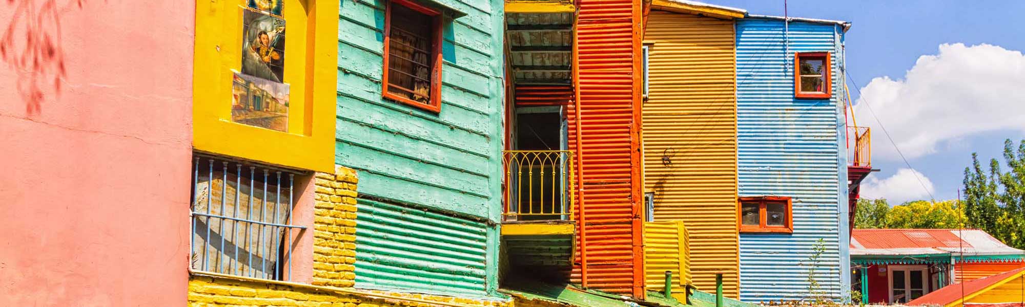 A vibrant row of traditionally painted corrugated metal houses in bright primary colors including pink, yellow, green, red, and blue. These iconic structures feature characteristic architectural details with shuttered windows, metal railings, and corrugated siding typical of the historic La Boca neighborhood.