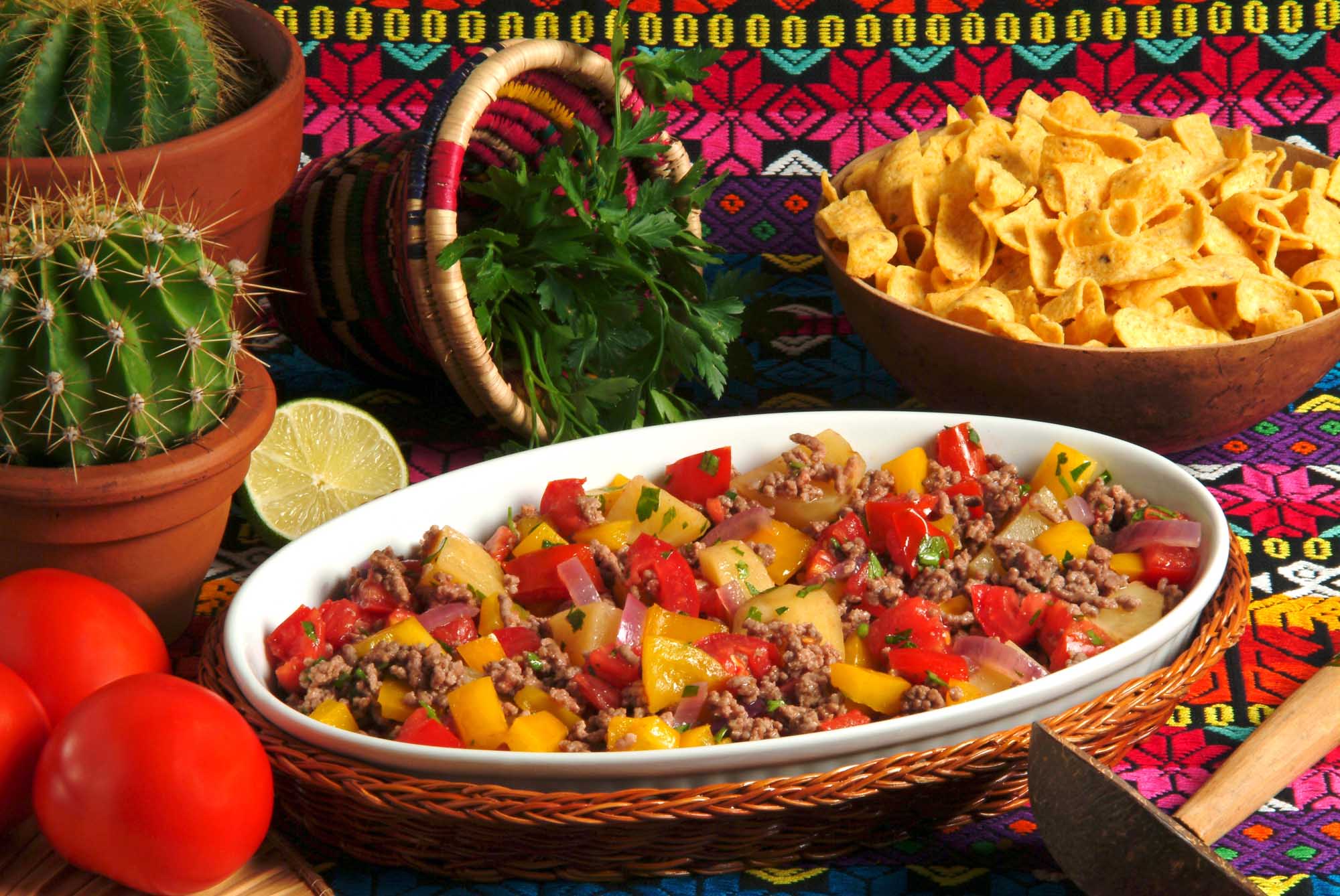 A vibrant presentation of Cuban picadillo habanero served in a white ceramic bowl, featuring seasoned ground meat with diced potatoes, red and yellow peppers, and fresh cilantro garnish. The dish is artfully arranged on a colorful woven placemat alongside complementary items including fresh lime, a decorative ceramic pitcher, potted cactus, crispy tortilla chips, and fresh tomatoes, creating an authentic and appetizing Cuban culinary scene.