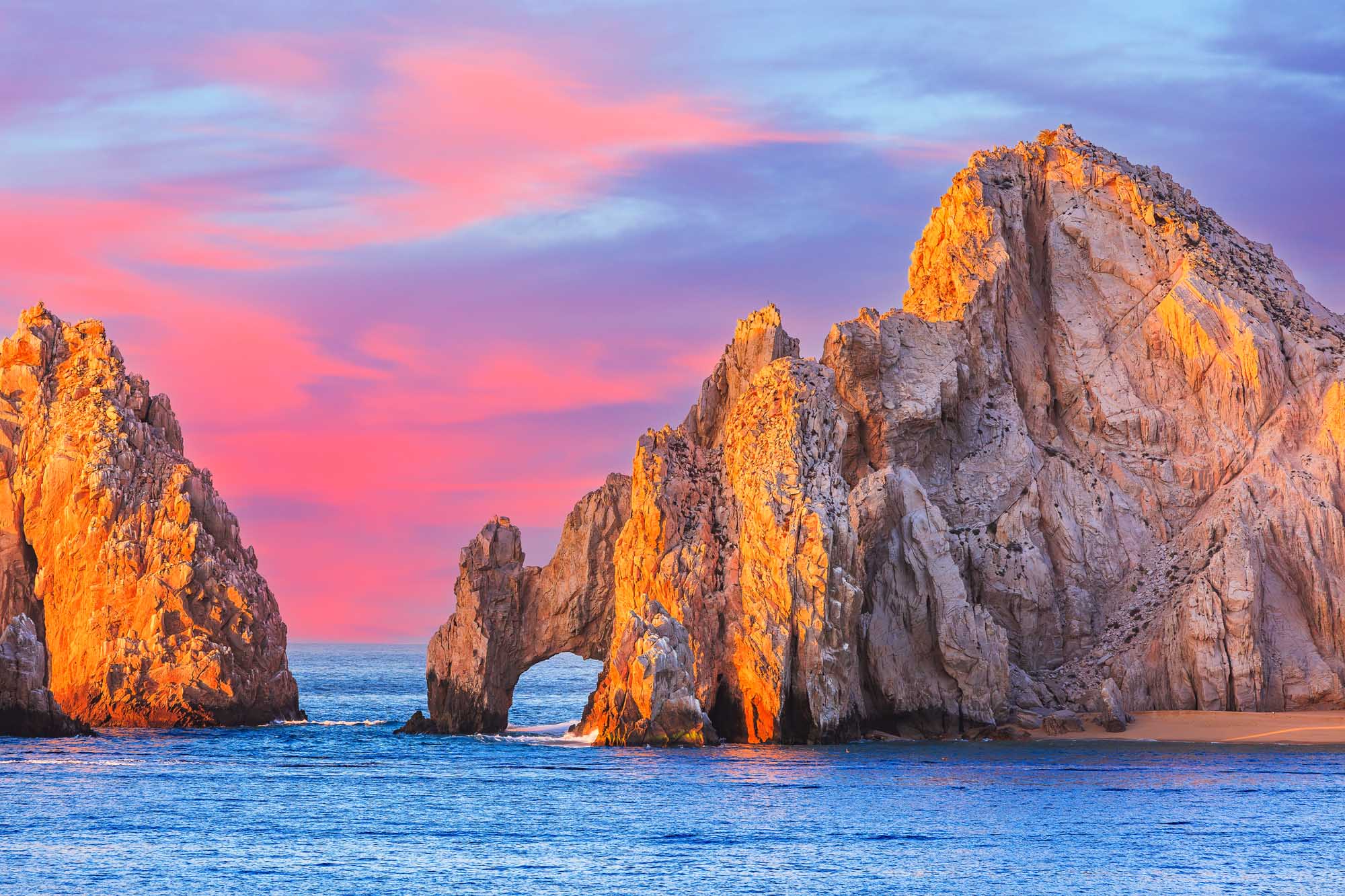 The iconic El Arco (The Arch) rock formation at Cabo San Lucas is bathed in golden sunset light, with dramatic granite cliffs rising from the turquoise Pacific waters.