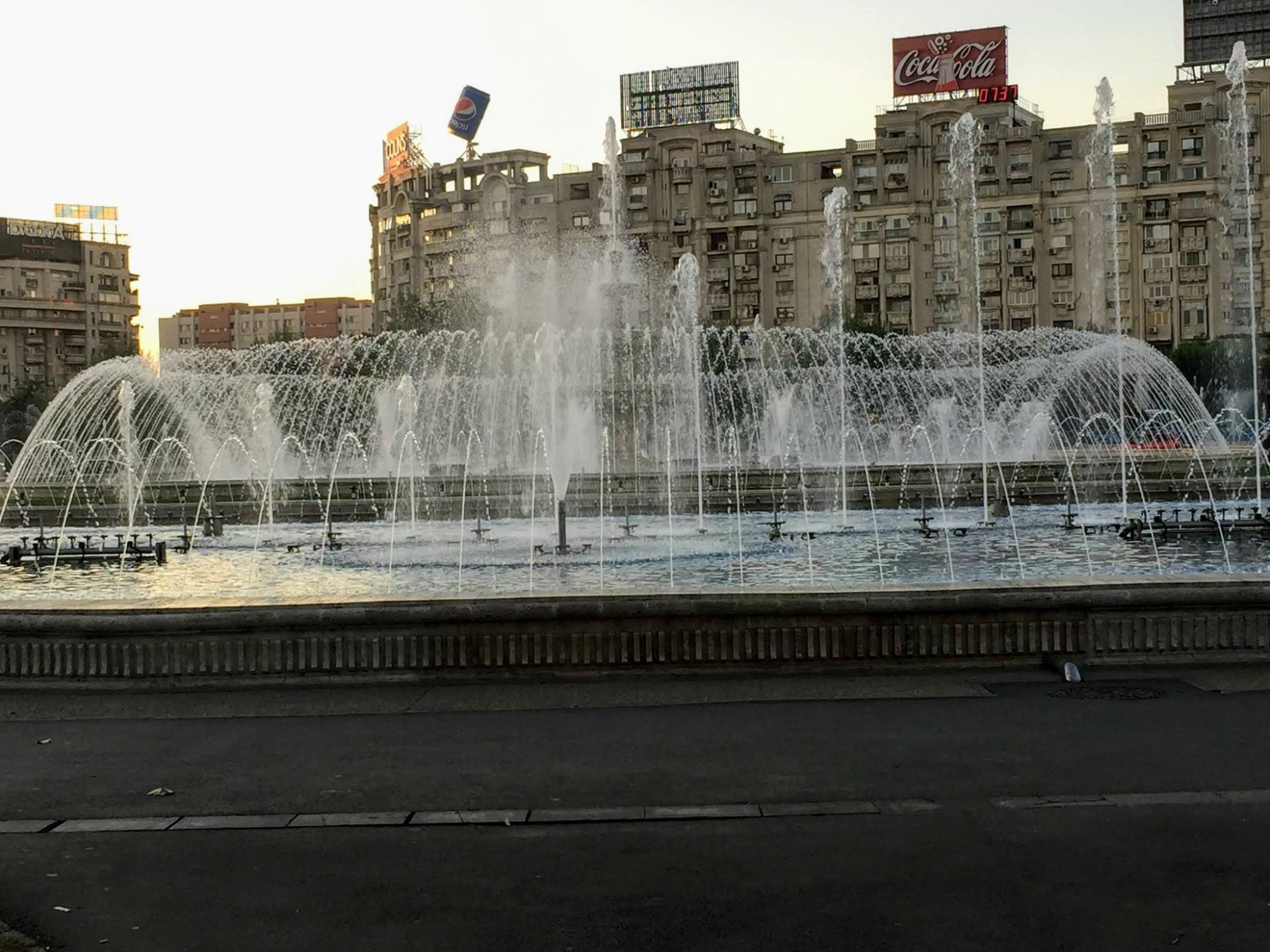 A spectacular large-scale fountain with multiple water jets and arching sprays in an urban plaza, featuring Soviet-era apartment buildings in the background and prominent vintage signage including a Coca-Cola advertisement on the roofline. The architectural style and urban design are characteristic of mid-20th century Eastern European cities.