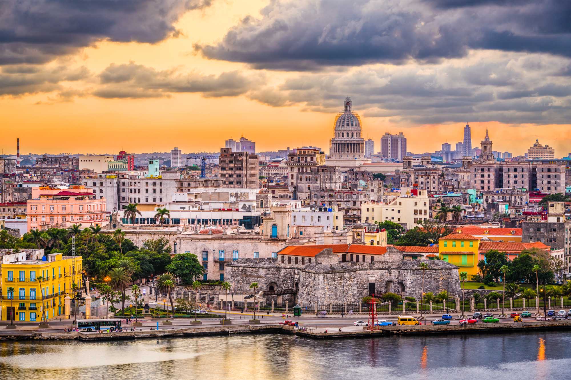 A stunning panoramic view of Havana, Cuba's downtown skyline at dusk, featuring the iconic Capitol building with its distinctive dome, colorful colonial architecture, palm trees, and the waterfront promenade (Malecon) along the harbor. The dramatic golden sky with storm clouds creates a striking backdrop for this vibrant Caribbean city.