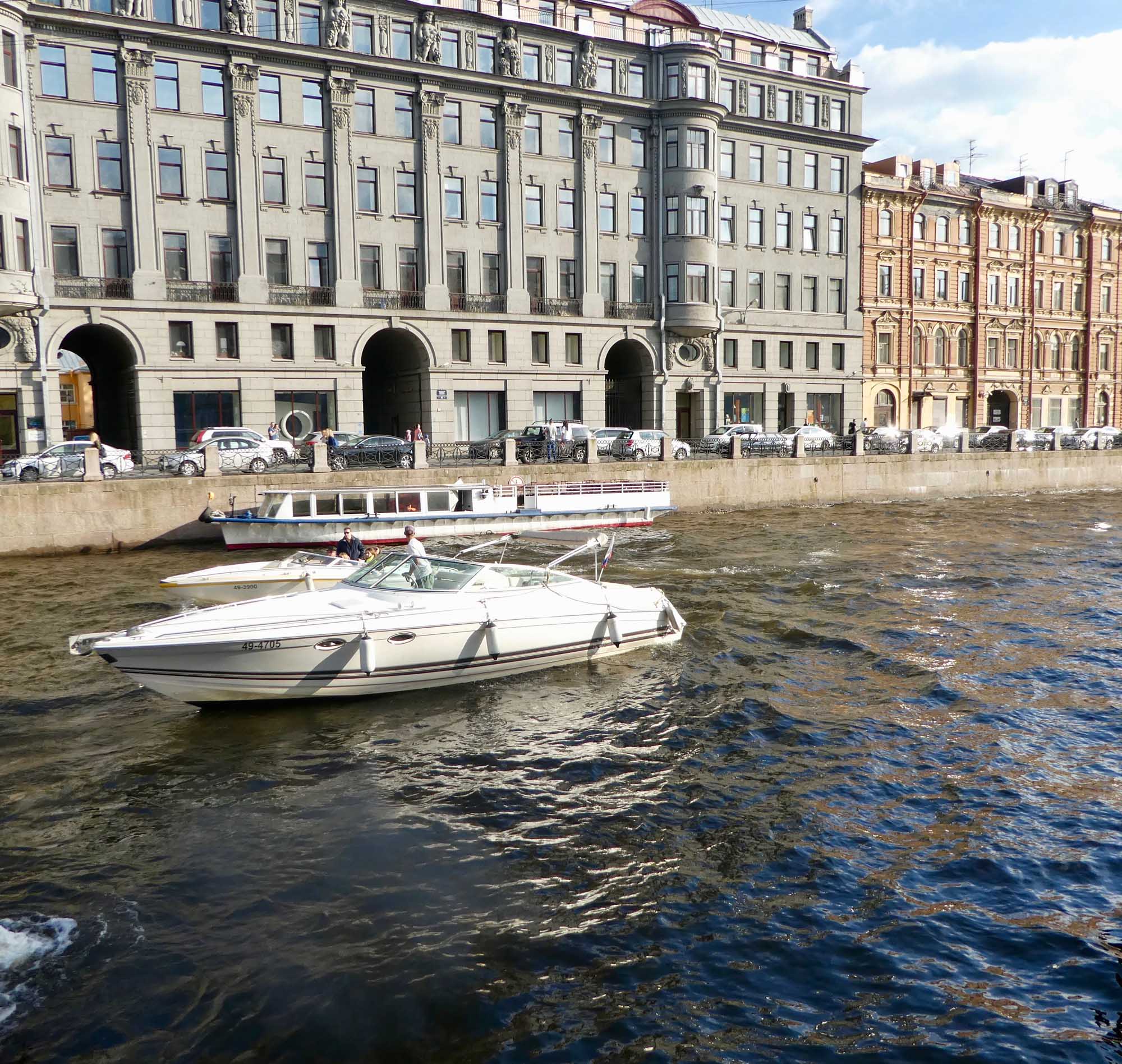 A picturesque waterfront scene featuring elegant historic European architecture with classical stone buildings, ornate columns, and mansard roofs lining the quay. White and blue leisure boats are moored in the calm water, with a passenger tour boat visible along the embankment.