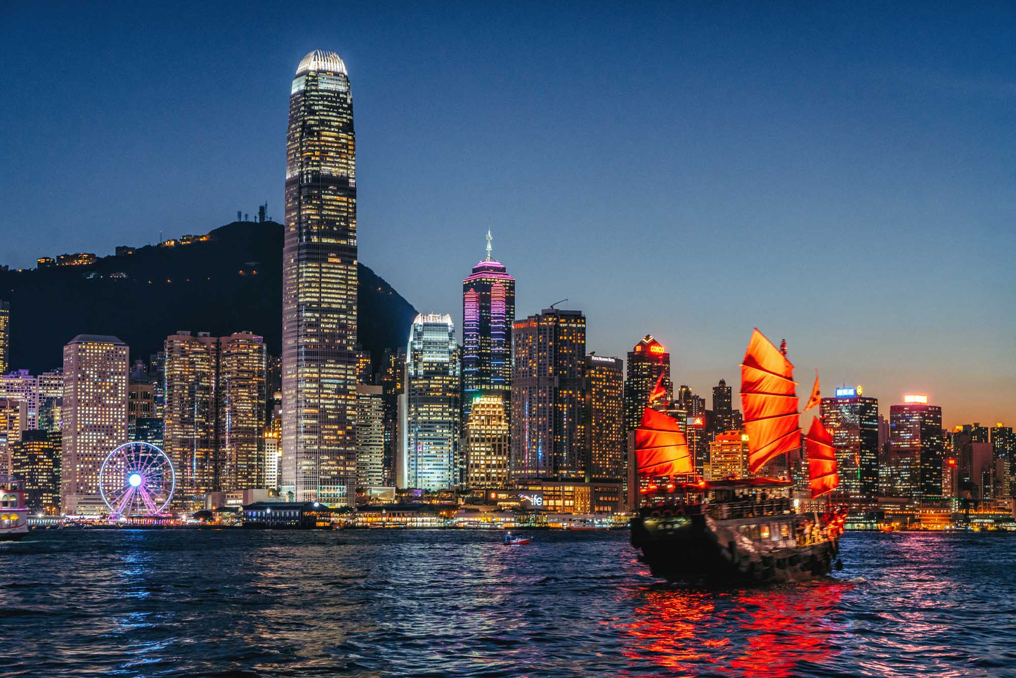 A stunning twilight view of Hong Kong's illuminated skyline featuring iconic skyscrapers including the International Finance Centre, with a traditional red-sailed junk boat prominently positioned in Victoria Harbour. The scene captures the contrast between modern architecture and historic maritime culture, with the Star Ferry Wheel and cityscape lights reflected in the water.