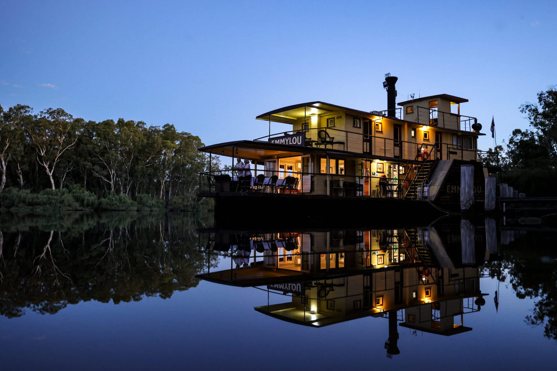 A striking nighttime image of the PS Emmylou, a heritage riverboat, beautifully lit against a twilight sky and mirrored perfectly in the still water below. The vessel is moored on a peaceful river lined with eucalyptus trees, creating a serene and atmospheric scene of river cruise tourism.