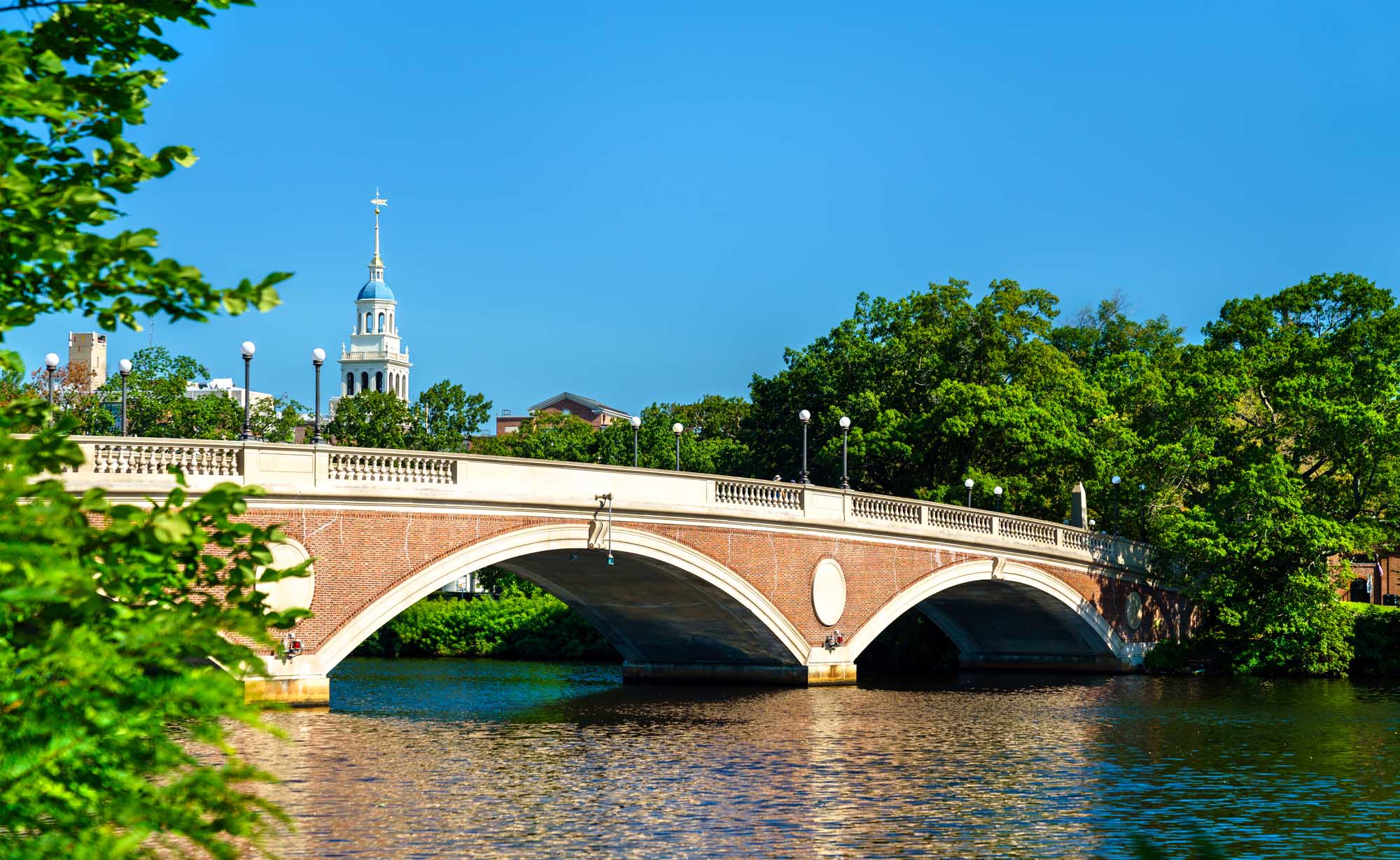 The iconic John W. Weeks Memorial Footbridge spans the Charles River between Boston and Cambridge, Massachusetts, featuring distinctive red brick arches and white stone balustrades. A historic church steeple is visible in the background under clear blue skies, with lush green trees framing the scenic riverscape.