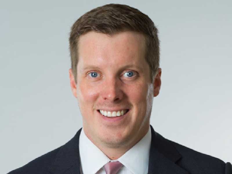 A professional portrait photograph of a man in business attire, wearing a dark navy blazer, white dress shirt, and mauve/pink tie, smiling warmly at the camera against a neutral light gray background.