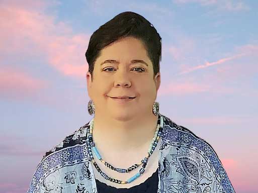 A professional headshot of a person wearing a patterned light blue jacket over a dark top, accented with beaded necklaces, photographed against a beautiful gradient sky featuring pink and blue sunset hues.