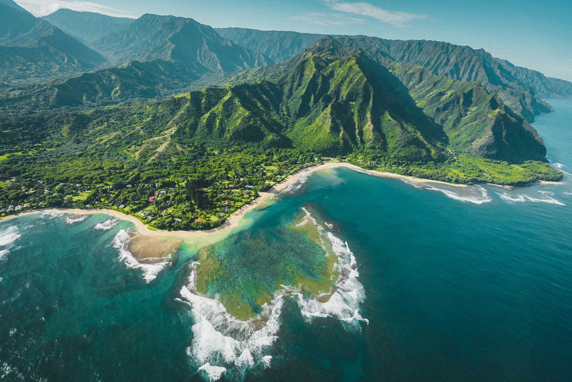 An aerial photograph of the lush Kalalau Valley on Kauai's dramatic Na Pali Coast, featuring verdant cliffs, a pristine sandy beach, turquoise waters, and white-capped ocean waves.