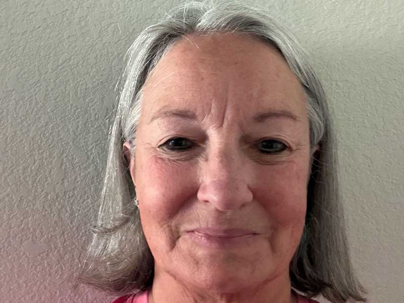A close-up portrait photograph of a middle-aged woman with shoulder-length gray hair wearing a pink/magenta colored top, photographed against a neutral beige textured wall. The image appears to be a personal or professional headshot taken indoors with natural lighting.