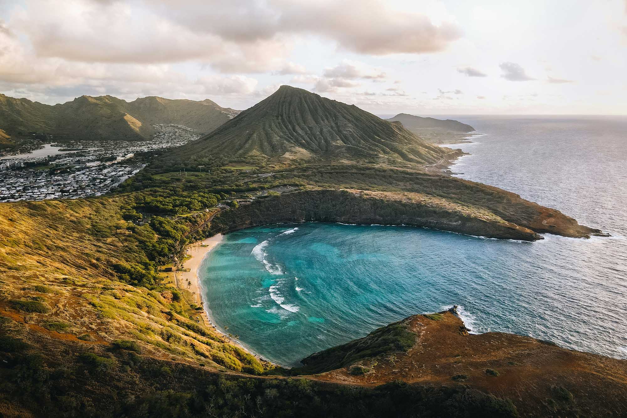 A stunning aerial view of Koko Crater and Koko Head in Hawaii, featuring a pristine turquoise bay with white sand beach nestled between dramatic volcanic ridges, with the iconic conical peak rising prominently in the background and a coastal town visible along the shoreline.