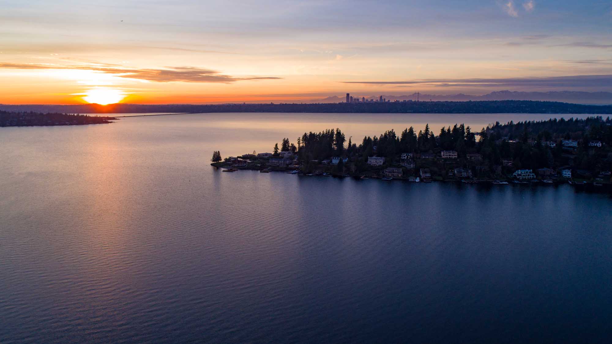 A breathtaking sunset view over Lake Washington, featuring a residential waterfront community with dense evergreen forests in the foreground and the Seattle skyline silhouetted against a golden and purple sky across the water.