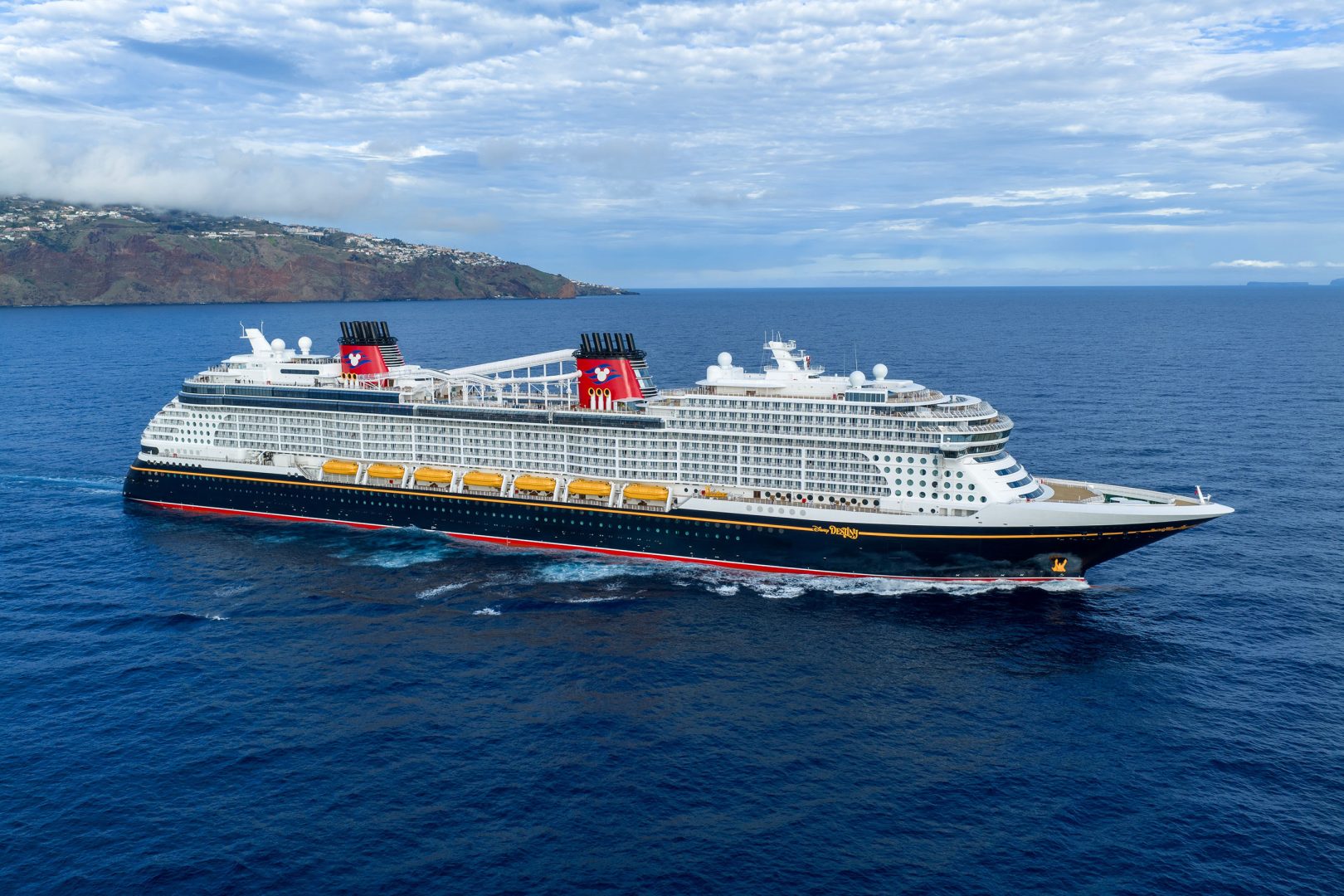 A massive modern cruise ship with distinctive red funnels featuring Mickey Mouse imagery is anchored in brilliant blue waters off a volcanic island with terraced settlements.