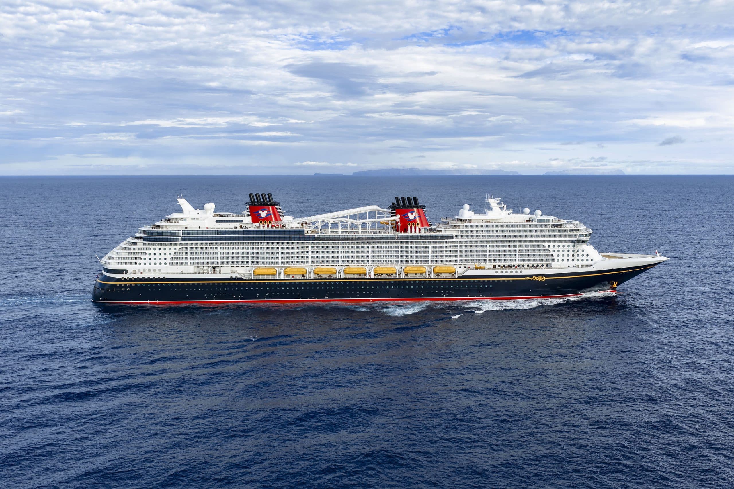 A large Disney cruise ship at sea with mountains in the background under clear skies.