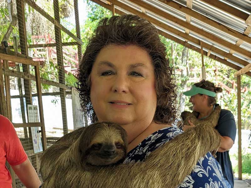 A woman smiles while holding a three-toed sloth in her arms at what appears to be an outdoor wildlife sanctuary or zoo facility. The setting features wooden structures with metal mesh enclosures and lush green vegetation, indicating an educational or interactive animal experience venue.
