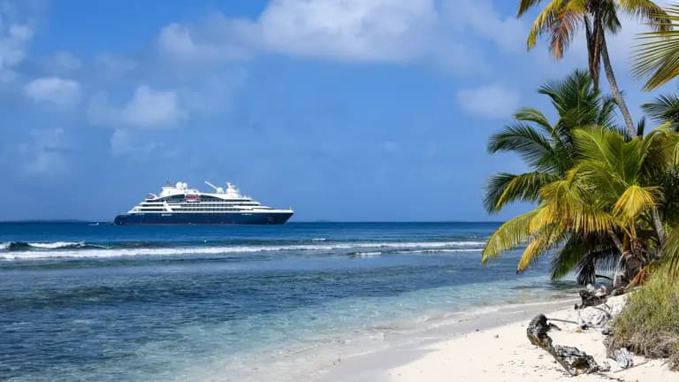 A large modern cruise ship anchors in clear turquoise waters off a pristine tropical beach lined with palm trees, exemplifying a classic Caribbean or Central American cruise destination with white sand, calm waters, and lush vegetation.