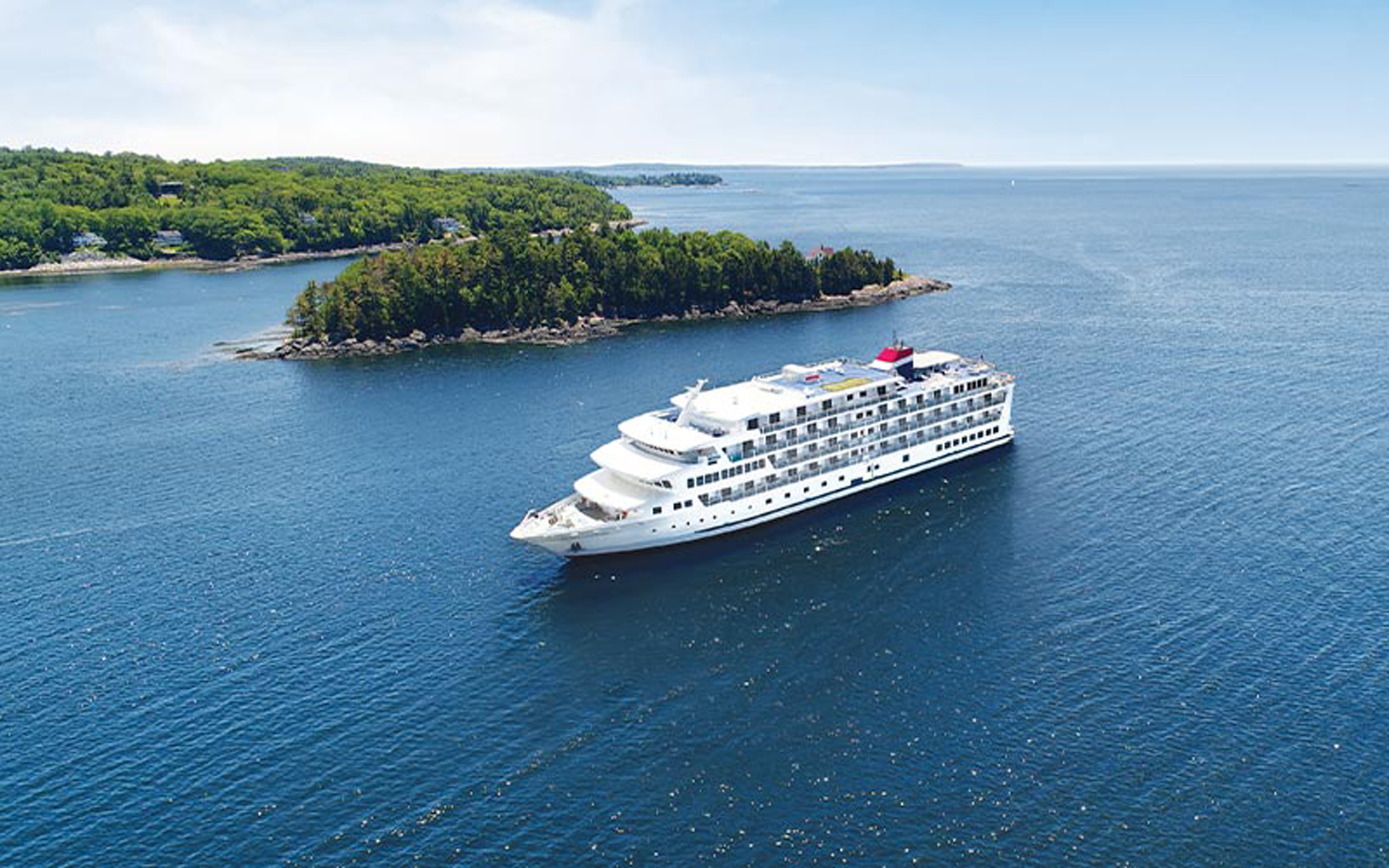 A large, modern cruise ship with distinctive white hull and red funnel anchors in the calm blue waters of Puget Sound, with a forested peninsula dotted with residential homes visible in the background.