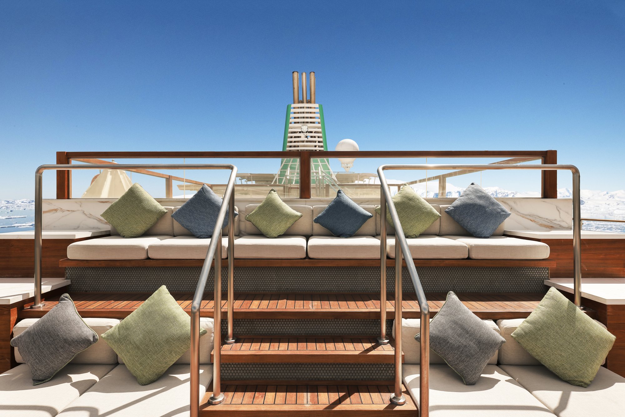 An elegant outdoor deck lounge area featuring modern seating with cream cushions and green accent pillows, arranged on wooden decking with metal frame structures. A distinctive funnel structure with three gold stacks rises prominently in the background, with snow-covered mountains and icy waters visible beyond the protective railings, suggesting an Arctic or polar cruise destination.