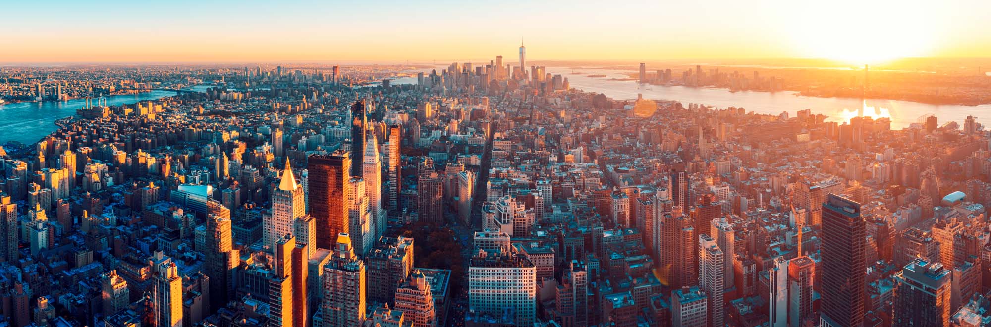 An expansive aerial panoramic view of Manhattan captured during golden hour, showcasing the iconic skyline with illuminated skyscrapers, the East River, Hudson River, and surrounding boroughs bathed in warm sunset light.